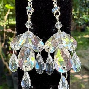 Puede incluir: Pendientes colgantes con detalles claros e iridiscentes en forma de lágrima y pétalo de cristal. Los pendientes tienen un engaste de metal plateado y se muestran sobre un fondo negro. Estos elegantes pendientes son adecuados para ocasiones especiales.