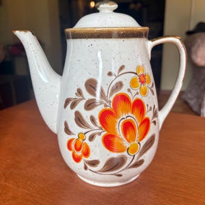 May include: White ceramic teapot with a lid, handle, and spout. The teapot is decorated with a floral design in orange, yellow, and brown. The rim and lid are trimmed with a brown speckled band. A vintage kitchenware item.