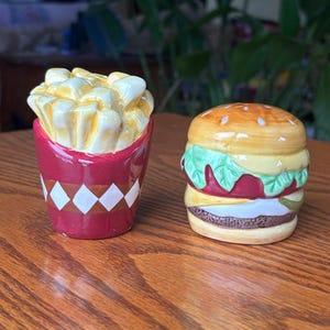 Peut inclure: Deux salières et poivrières en céramique en forme de frites dans un récipient rouge à motifs losanges blancs et d'un hamburger avec un pain aux graines de sésame, de la laitue et un steak.