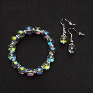 Könnte beinhalten: Ein Set aus Ohrringen und einem Armband aus irisierendem, klarem Glas. Die Ohrringe sind silberfarben mit einem klaren, tropfenförmigen Glasstein. Das Armband besteht aus klaren Glasperlen mit irisierenden Regenbogenfarben.
