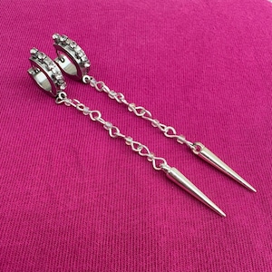 May include: Silver-coloured dangle earrings with a hoop design encrusted with small, clear rhinestones. The earrings feature a chain of small links and beads, culminating in a pointed, cone-shaped pendant. The earrings are displayed against a pink background.