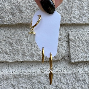 May include: Three gold-toned hoop earrings attached to a white display card. One hoop features a clear, hexagonal crystal pendant, one is a plain hoop, and the third hoop features a gold-toned, pointed, cone-shaped metal charm hanging from it.