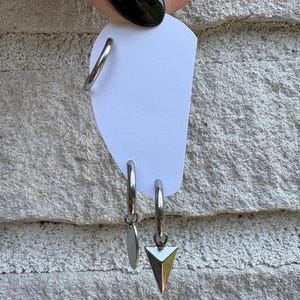 Puede incluir: Un conjunto de tres pendientes de aro de metal plateado sujetos a una tarjeta blanca. Un aro es liso, otro presenta un pequeño dije de disco circular y el tercer aro incluye un dije de punta triangular tridimensional colgando de la base.