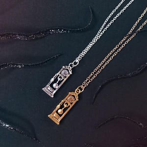 May include: Two necklaces featuring clock-shaped pendants. One is silver-coloured, the other gold-coloured. The pendants have detailed clock faces and pendulum designs. The chains are fine, and the background is black.