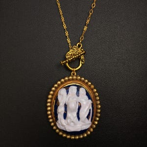 May include: Gold chain necklace with a blue and white cameo pendant featuring three women in a relief design. The pendant is surrounded by a gold beaded border.