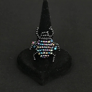 Peut inclure: Une bague en perles noires avec un motif de scarabée multicolore. Le scarabée est composé de perles bleues, vertes et violettes irisées.