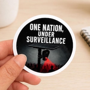 May include: A circular sticker featuring a black background with the white text ONE NATION, UNDER SURVEILLANCE. Below the text is a dark silhouette of a crowd under a pole-mounted camera emitting a red light beam directed at the people below.