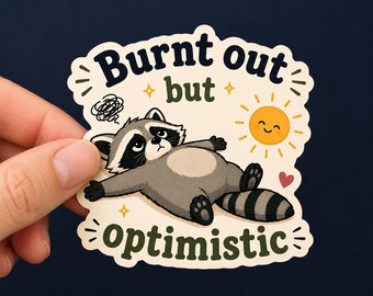 Può includere: Un adesivo fustellato raffigurante un procione grigio e nero sdraiato sulla schiena con un'espressione stanca. Sopra il procione, il testo recita Burnt out but, e sotto optimistic. Sono inclusi un sole giallo sorridente e un piccolo cuore rosso.