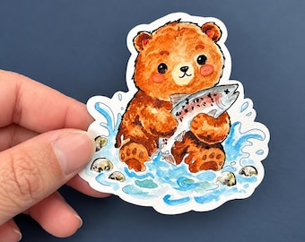 Può includere: Un adesivo fustellato con un'illustrazione ad acquerello di un cucciolo di orso bruno con le guance rosate che tiene in mano un pesce argentato. L'orso è seduto in acqua blu che schizza con piccole pietre grigie sparse alla base.