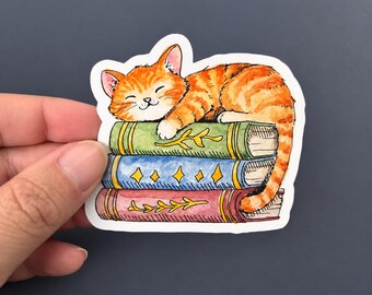 Può includere: Un adesivo fustellato raffigurante un gatto soriano arancione che dorme su una pila di tre libri. Il libro in alto è verde con motivi a foglie dorate, quello centrale è blu con stelle dorate e quello in basso è rosso con foglie dorate.