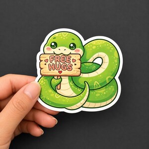 May include: A die-cut vinyl sticker featuring a bright green snake with a light cream underbelly, coiled in a circle. The snake holds a small, light brown wooden sign with the text FREE HUGS in dark pink letters, accented by small pink hearts.