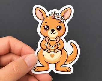 Può includere: Un adesivo fustellato raffigurante un canguro arancione in stile cartone animato con una corona di fiori sulla testa. Il canguro ha un piccolo cucciolo che spunta dal marsupio, contrassegnato da un piccolo cuore marrone. Il design ha un bordo bianco.