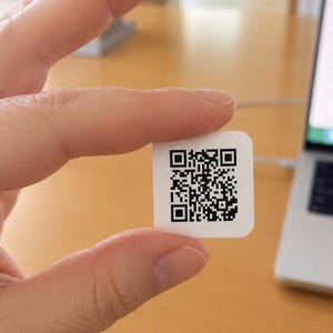 May include: A small, square, white sticker featuring a black and white QR code. The sticker has rounded corners and a smooth, matte finish, displaying a complex, high-contrast digital pattern designed for scanning and data retrieval purposes.
