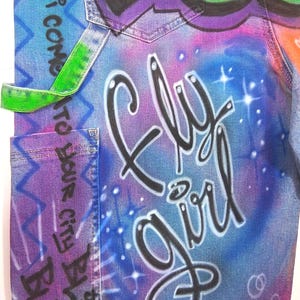 May include: Close-up of denim fabric with graffiti art. The design features the words "Fly Girl" in black script, set against a blue and purple gradient background. Green and purple graffiti art is at the top.
