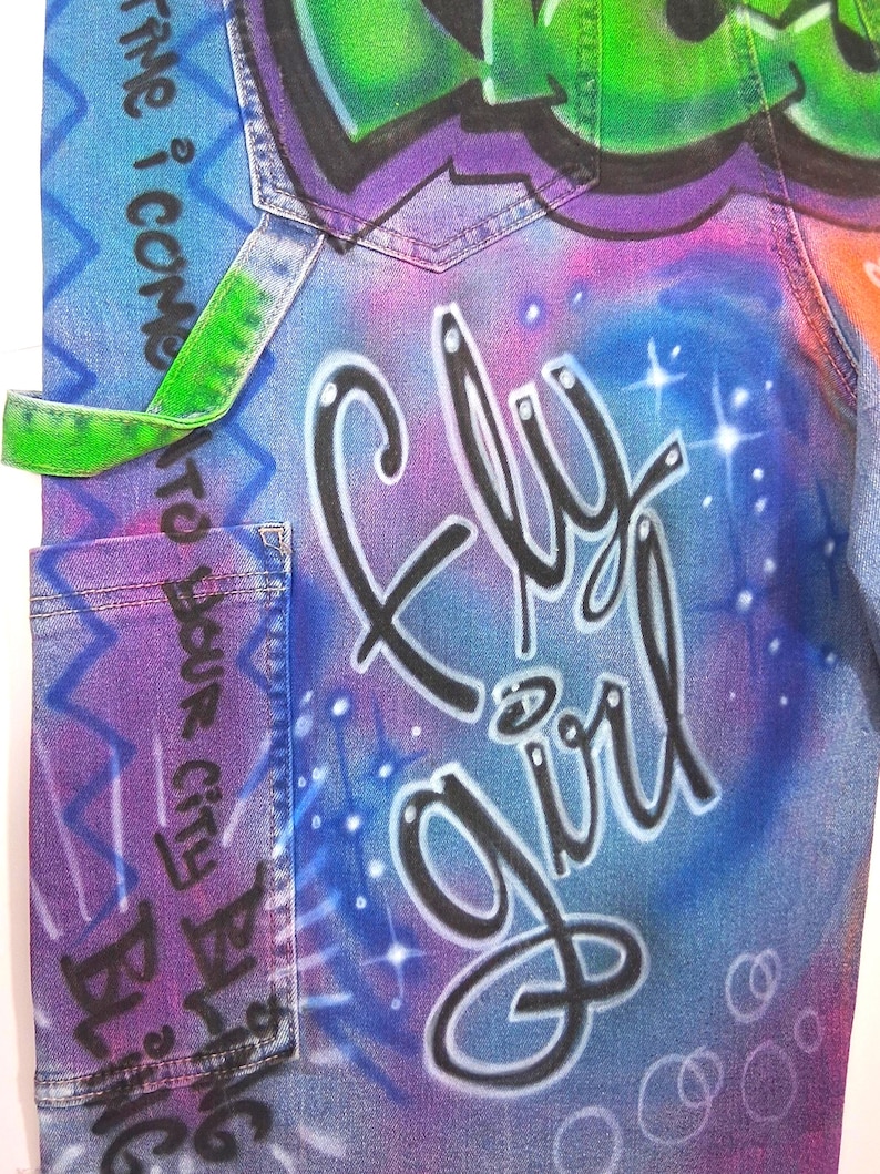 May include: Close-up of denim fabric with graffiti art. The design features the words "Fly Girl" in black script, set against a blue and purple gradient background. Green and purple graffiti art is at the top.