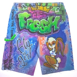 May include: Denim shorts with airbrushed graffiti art. The design features the word "FRESH" in green, "Fly Girl" in cursive, and a cartoon character. The shorts have a distressed hem and a green accent on the pocket.