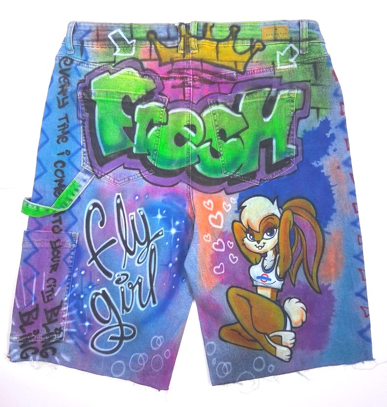 May include: Denim shorts with airbrushed graffiti art. The design features the word "FRESH" in green, "Fly Girl" in cursive, and a cartoon character. The shorts have a distressed hem and a green accent on the pocket.