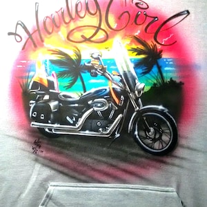 May include: A gray hoodie featuring an airbrushed design of a black motorcycle with a windshield, set against a tropical sunset scene with palm trees and the text "Harley Girl" in cursive.