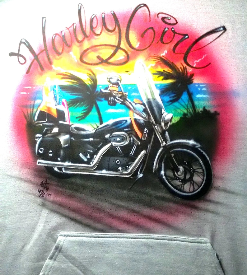May include: A gray hoodie featuring an airbrushed design of a black motorcycle with a windshield, set against a tropical sunset scene with palm trees and the text "Harley Girl" in cursive.