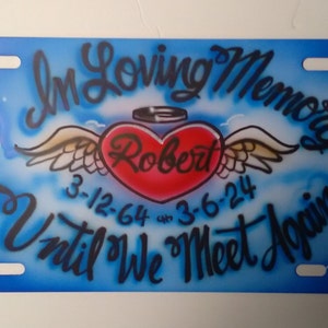 May include: A blue and white memorial license plate with a red heart with wings and the text "In Loving Memory Robert 3-12-64 -- 3-6-24 Until We Meet Again".