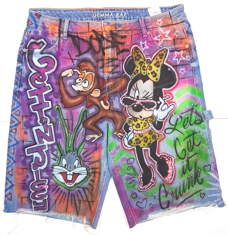 May include: Denim shorts with colorful graffiti art. The shorts feature cartoon characters, including a monkey, Bugs Bunny, and Minnie Mouse. The shorts have a distressed hem and the text "Let's Get it Crunk".