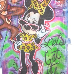 May include: Denim jeans with a vibrant, colorful design featuring Minnie Mouse in a leopard print dress and bow, wearing sunglasses. The jeans have graffiti-style lettering that says "Lets Get it Crunk" and a cartoon monkey.