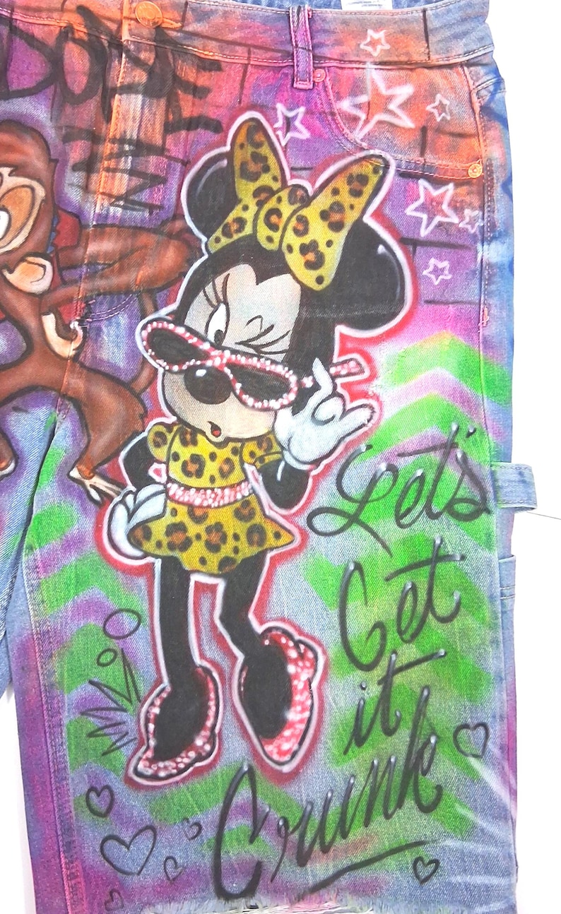 May include: Denim jeans with a vibrant, colorful design featuring Minnie Mouse in a leopard print dress and bow, wearing sunglasses. The jeans have graffiti-style lettering that says "Lets Get it Crunk" and a cartoon monkey.