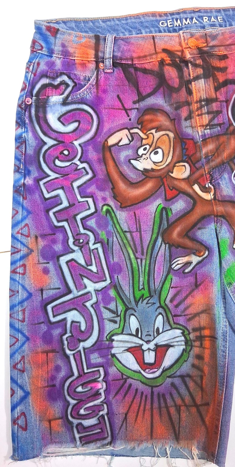 May include: Denim shorts with a colorful graffiti design featuring cartoon characters and the text "Goth N' Love". The shorts have a distressed hem and the brand name "GEMMA RAE" is visible.