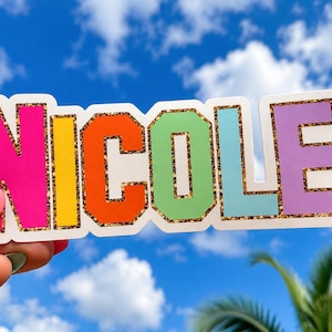 May include: A die-cut sticker featuring the name NICOLE in bold, block capital letters. Each letter is a different vibrant color, including hot pink, yellow, orange, light green, light blue, and purple, all outlined with a gold glitter border.