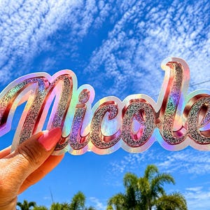 May include: A custom name sticker featuring the name Nicole in a cursive script font. The letters have a textured silver glitter finish with a metallic rose gold outline and a holographic border that reflects light in various rainbow colors.