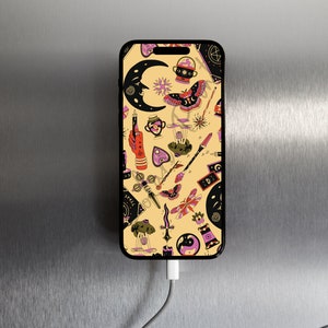 Puede incluir: Un teléfono inteligente negro con una funda de fondo amarillo que presenta un patrón de símbolos ocultos negros y rosas, incluyendo una luna creciente, un pentagrama, una mano sosteniendo una vela, una mariposa y una daga. El teléfono está conectado a un cable de carga blanco.