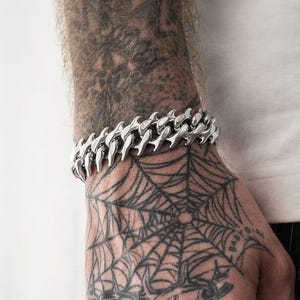 以下が含まれることがあります： ユニークでとがったデザインのシルバーチェーンブレスレットが手首に着用されています。手には、いくつかのシルバーリングと、クモの巣のデザインを含む複雑なタトゥーもあります。その人は白いシャツを着ています。