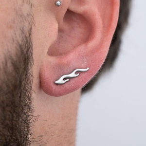 Puede incluir: Un pendiente plateado con forma de llama se lleva en la oreja. El pendiente tiene un diseño curvo que imita la forma de una llama. El pendiente es de estilo stud y está hecho de un metal brillante. El fondo es blanco liso.