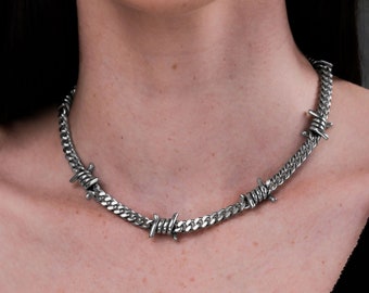 May include: A silver-colored necklace featuring a curb chain design with barbed wire accents. The necklace is worn around the neck and is made of a shiny metal material. The barbed wire adds a unique, edgy aesthetic to the jewelry.