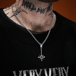 May include: A silver-colored pendant necklace with a cross-like design. The necklace hangs from a chain and is worn over a black mesh shirt. The word "VERY" is visible on the shirt.