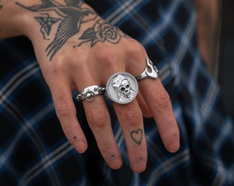May include: A hand wearing three silver rings. The ring on the middle finger features a skull design. The ring on the index finger has a small circle with a ring attached. The ring on the little finger has a flame design.