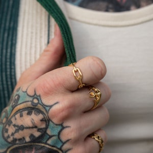 May include: Two gold rings on a hand with a tattoo. One ring is a chain link design and the other is a stylized wrench.
