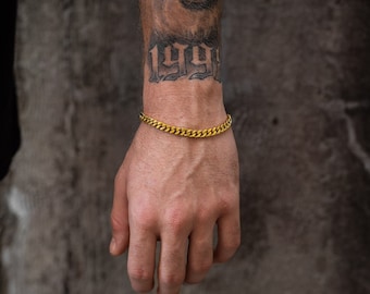 May include: A gold-colored curb chain bracelet. The bracelet is worn on a wrist with a tattoo of the number "1991". The bracelet has a series of interlocking links, creating a textured surface. The bracelet is a simple yet stylish accessory.