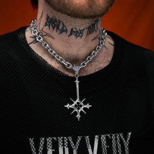 May include: Silver-toned necklace with a pendant featuring a stylized cross design. The necklace has a chunky chain and is worn over a black mesh shirt. The pendant has a starburst design. The shirt has the words "VERY VERY" printed on it.