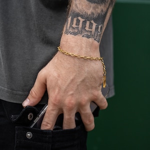 Puede incluir: Un brazalete de cadena dorada con un pequeño dije colgante. El brazalete se lleva en la muñeca de una persona con un tatuaje del año 1998.
