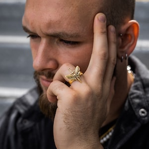 May include: A gold ring featuring a detailed design of a moth with a dagger piercing its body.