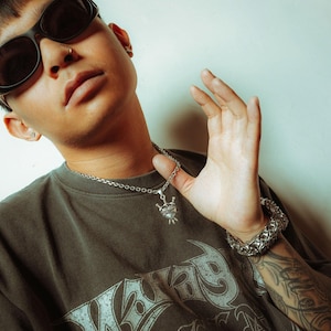 Silver heart pendant with 3 swords going through it and wrapped in chain on male model holding the chain up with glasses