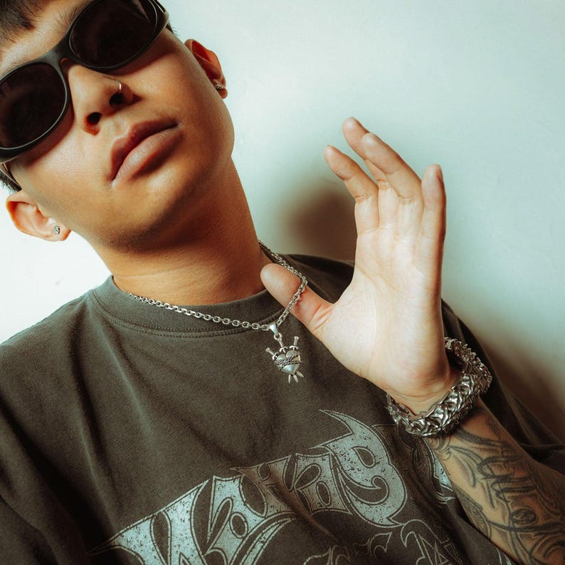 Silver heart pendant with 3 swords going through it and wrapped in chain on male model holding the chain up with glasses