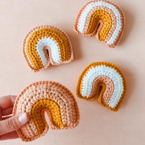 May include: Four small crocheted rainbows in varying shades of peach, mustard yellow, and white.  Each rainbow features three distinct color bands, creating a layered effect. The rainbows are crafted from a textured yarn, giving them a soft, handmade appearance. These charming, miniature rainbows are perfect for nursery decor or as small, whimsical gifts.