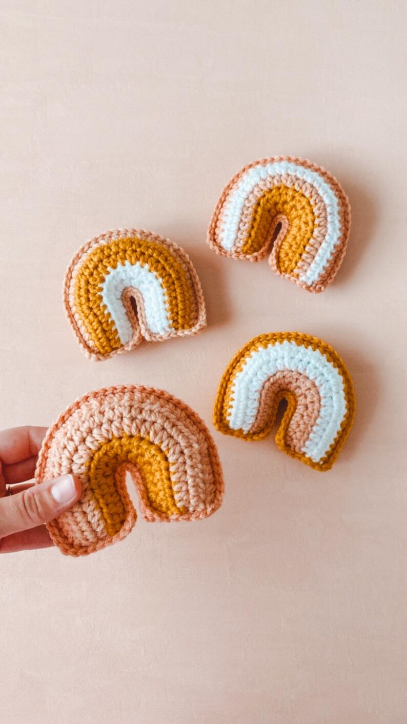 May include: Four small crocheted rainbows in varying shades of peach, mustard yellow, and white.  Each rainbow features three distinct color bands, creating a layered effect. The rainbows are crafted from a textured yarn, giving them a soft, handmade appearance. These charming, miniature rainbows are perfect for nursery decor or as small, whimsical gifts.