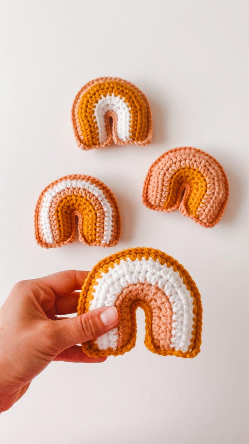 May include: Four small crocheted rainbows in varying sizes. Each rainbow features three color bands: mustard yellow, off-white, and dusty rose. The rainbows are crafted from soft yarn, creating a textured appearance. These handmade items are perfect for nursery decor or as small, charming gifts.