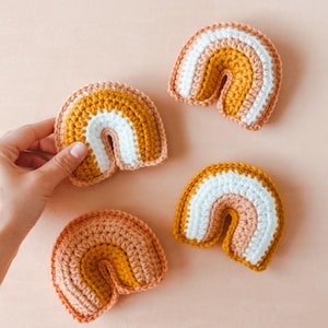 May include: Four small crocheted rainbows in peach, mustard yellow, and white.  The rainbows are arranged on a light peach background. Each rainbow is made of three color bands and has a slightly different size and shade variation. These handmade crocheted rainbows are perfect for nursery decor or as a baby shower gift.