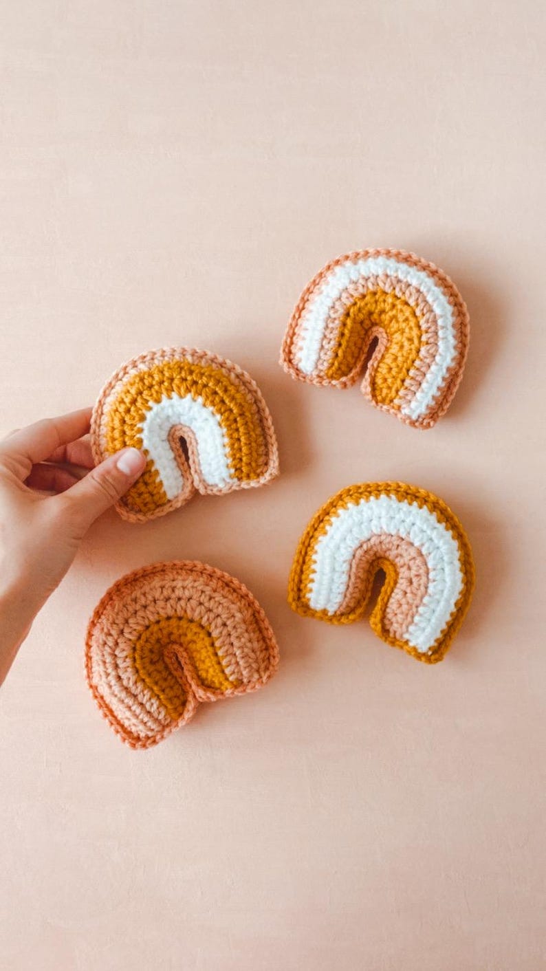 May include: Four small crocheted rainbows in peach, mustard yellow, and white.  The rainbows are arranged on a light peach background. Each rainbow is made of three color bands and has a slightly different size and shade variation. These handmade crocheted rainbows are perfect for nursery decor or as a baby shower gift.