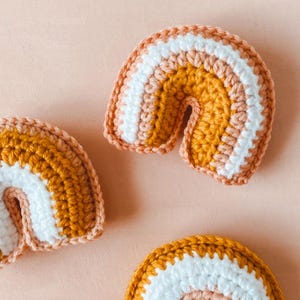 May include: Three small crocheted rainbows in varying sizes. Each rainbow features stripes of mustard yellow, peach, and white yarn. The rainbows are arranged on a light peach background. These handmade rainbow decorations are perfect for nurseries or children's rooms.