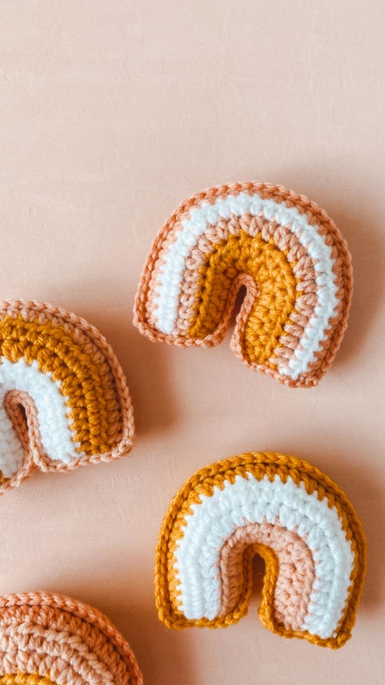 May include: Three small crocheted rainbows in varying sizes. Each rainbow features stripes of mustard yellow, peach, and white yarn. The rainbows are arranged on a light peach background. These handmade rainbow decorations are perfect for nurseries or children's rooms.